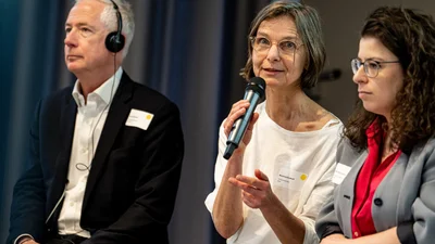 ein Mann und zwei Frauen, eine davon hält ein Mikro in der Hand und spricht