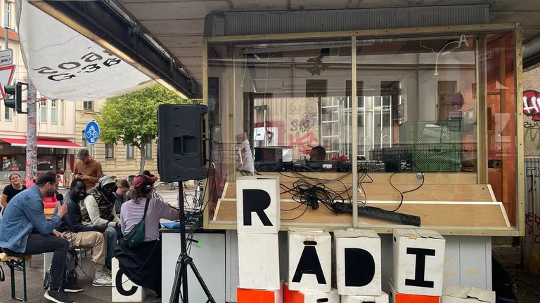 Small mobile radio booth with large letter blocks spelling »RADIO« and several people sitting on benches nearby.