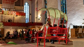 Zwei Personen sitzen auf einer roten Bank mit einem Rosenbogen ähnlichen Sonnendach in einem Kirchenraum mit Menschen im Hintergrund