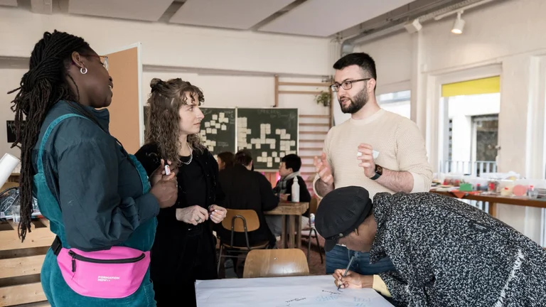 vier Personen überlegen gemeinsam und stehen um einen Tisch, auf dem ein Plakat liegt.