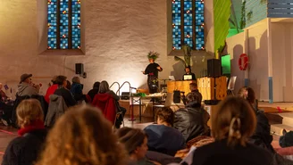 Person mit pflanzen ähnlichem Kopfschmuck steht vor Publikum und führt etwas auf