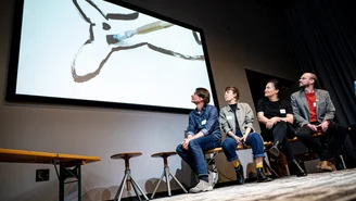 four people sit on the stage and look at the screen diagonally behind them, on which a drawing can be seen