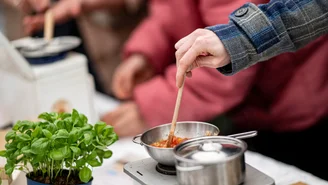 Close-up of a miniature hob with two miniature pots on it. A hand is stirring something in them with a wooden spoon. Next to it is a normal-sized basil pot