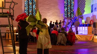 People wearing large flower costumes stand in a church in front of a group of seated and standing people