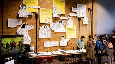 An einem langen Tisch stehen mehrere Personen für die Anmeldung. Dahinter an einer Macksteinwand hängen verschiedene Comics und Texte dazu