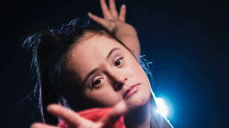A dancer in front of a dark background looks directly into the camera and ‘reaches’ for the viewer with her hand