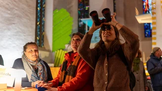 Drei Personen in einem Innenraum mit bunten Glasfenstern, eine Person hält ein Fernglas nach oben gerichtet