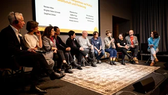 Eleven people are sitting on benches on the stage, one woman is holding a microphone and speaking. The others look towards her