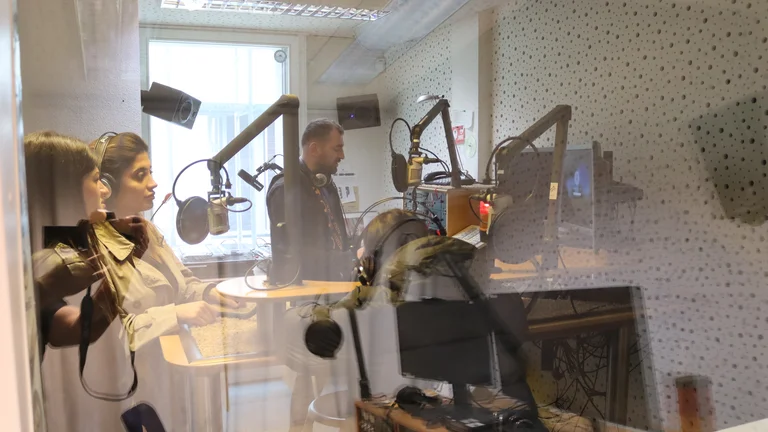 Zwei Personen stehen in einem Radiostudio mit Mikrofonen, Mischpult und Computern.