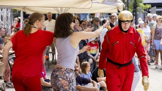 Two women stretch out their arms to press a button on the shoulder of a human dressed as a robot.