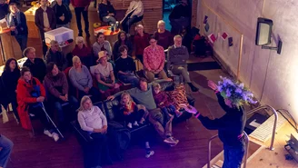 Person steht auf einer Bühne und spricht vor einem Publikum, das auf Stühlen und Sofas sitzt