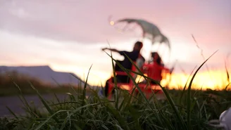 Verschwommene Silhouetten von zwei Personen, die auf einer Bank im Gras sitzen, im Hintergrund ein Sonnenuntergang.