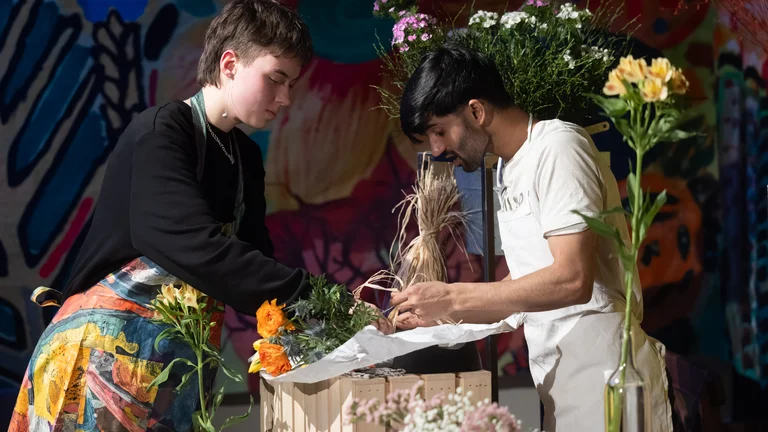Ein junger Mann und eine junge Frau haben Schürzen an und binden zusammen einen Blumenstrauß. Premiere des Theaterstücks Unsichtbare Wunden.