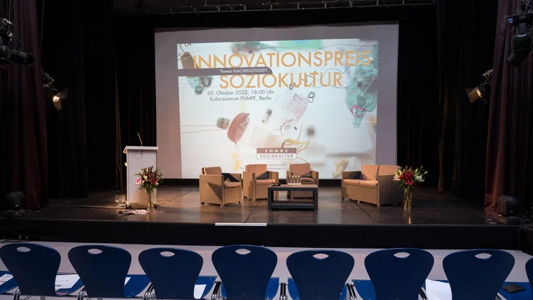 noch leere Stühle vor einer Bühne mit Sesseln. Dahinter eine Leinwand mit einer Präsentation mit der Aufschrift Innovationspreis Soziokultur