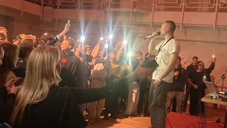 A young man is standing on a small stage, singing into a microphone, with an audience of young people in front of him.