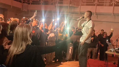 Ein junger Mann steht auf einer kleinen Bühne und singt in ein Mikrofon, vor im ist ein Publikum aus jungen Menschen.