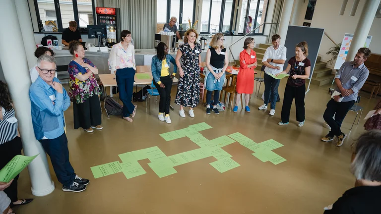 eine Gruppe von Menschen steht im Kreis, in der Mitte liegen verschiedene Zettel auf dem Boden