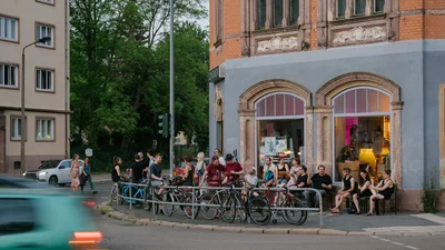 Außenansicht eine Veranstaltungsortes mit großen Fenstern, vor denen verschiedene Personen sitzen und sich unterhalten.