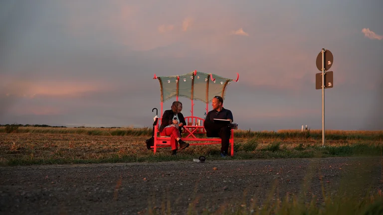 zwei Personen unterhalten sich sitzend auf einer Bank an einer Straße auf dem Land