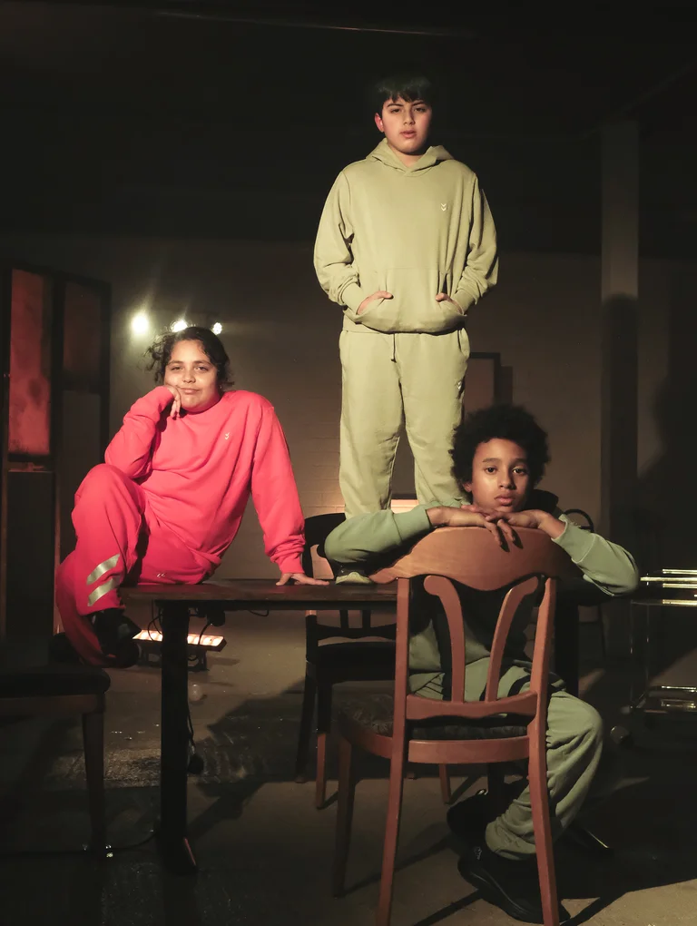 Drei Kinder stehen und sitzen auf einem Tisch und einem Stuhl und schauen in die Kamera.