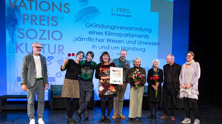 Neun Menschen auf der Bühne mit Blumensträußen und einer Urkunde in der Hand