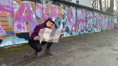 Eine junge Person in der Hocke vor einer bunten Graffiti-Wand hält eine aufgefaltete Karte in den Händen.
