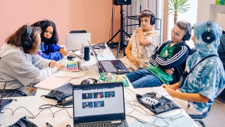A group of young people sit with headphones around a table on which various technical devices such as laptops, microphones and cables are lying