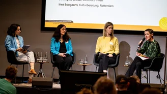 Three women sit on chairs on the stage and look at a fourth woman, who is also sitting and speaking with a microphone in her hand