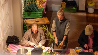 Drei Personen sitzen an einem Tisch und bereiten Gemüse zu, im Hintergrund Obst- und Gemüsekisten.