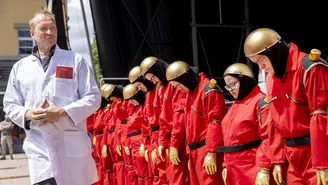 People dressed as robots stand in a row with their heads bowed. A man in a doctor's coat walks in front of them