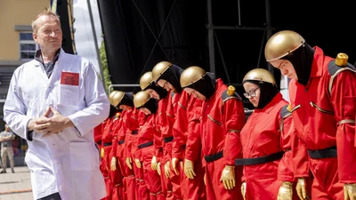 Als Roboter verkleidete Menschen stehen in einer Reihe mit geneigtem Kopf. Vor ihnen läuft ein Mann mit einem Arztkittel