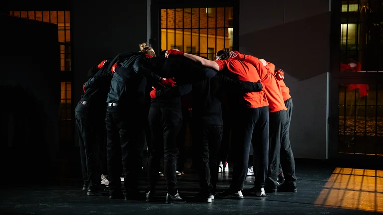 A group of young men put their arms around each other and form a circle while looking down. Windows closed with bars can be seen in the background.