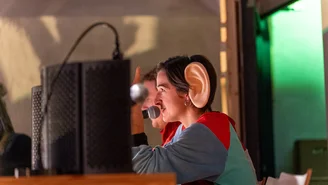 Person mit übergroßem, künstlichem Ohr sitzt an einem Tisch mit Mikrofon und Lautsprechern im Hintergrund